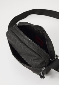 Sort tekstureret crossbody taske med to lynlåse, justerbar rem og rød "HUGO" label indeni hovedrum.