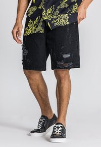 Pantalones cortos de mezclilla negra con detalles desgastados, dobladillos deshilachados y un corte relajado, combinados con zapatillas negras y blancas sobre un fondo claro.