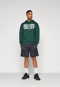 Felpa verde dei Boston Celtics con testo bianco, abbinata a pantaloni corti in mesh neri. Sneakers nere e calzini bianchi visibili. Abbigliamento sportivo semplice e casual.