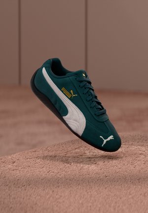 Baskets Puma couleur sarcelle avec bande latérale blanche, semelle noire, logo Puma doré et lacets flottant au-dessus d'une surface beige texturée.