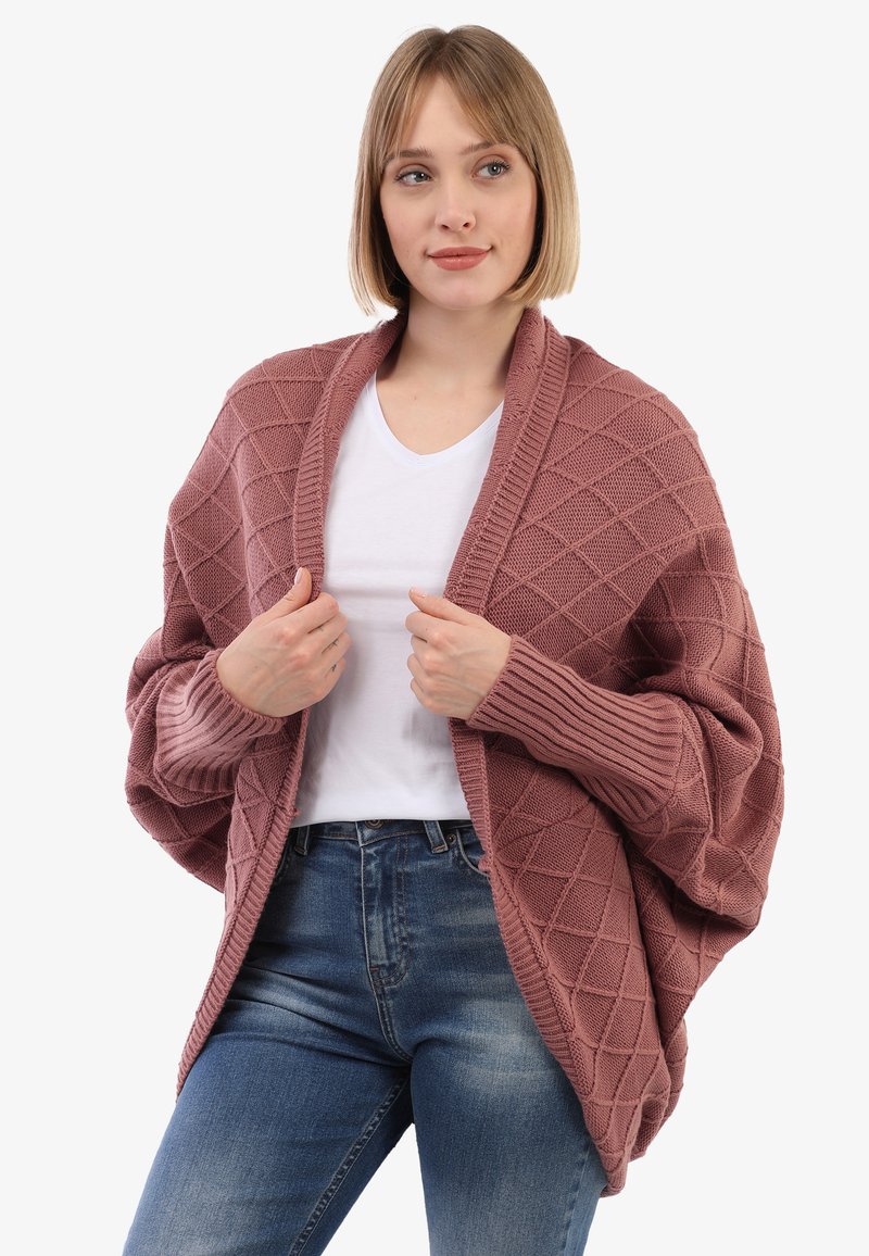Frau, die eine pinkfarbene, übergroße Strickjacke mit Rautenmuster über einem weißen T-Shirt und blauen Jeans trägt und die Kanten der Strickjacke hält.