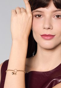 Złota bransoletka z łańcuszka z zawieszkami w kształcie serca i gwiazdy, o fakturowanym wzornictwie. Modelka nosi bordową bluzkę, prezentującą akcesorium na nadgarstku.