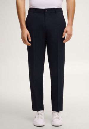 Mann trägt schlanke dunkelblaue Hose und weiße Turnschuhe, steht vor einem schlichten hellen Hintergrund.