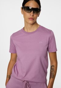 Lyselilla kortærmet t-shirt med rund halsudskæring og subtil logo detaljer. Modellen bærer mørke solbriller, som viser en glat stofstruktur.