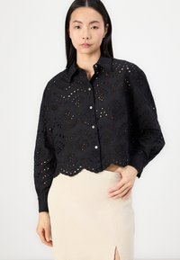 Black cropped shirt with intricate eyelet embroidery, scalloped hem, and button closure. Long sleeves with elastic cuffs.