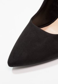Svart mocka sko med spetsig tå och låg skärning, med en jämn yta och en elegant silhuett, mot en vit bakgrund.