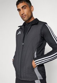 Chaleco acolchado negro con un cuello alto, cierre frontal de cremallera y bolsillos laterales. Presenta detalles en gris y tres rayas blancas en las mangas.