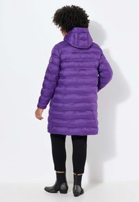 Lila Steppjacke mit Kapuze, horizontalen Steppnähten, einem Logo auf dem Ärmel und einem langen Schnitt. Getragen mit schwarzen Hosen und besetzten Stiefeln.