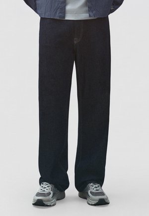 Modèle portant un jean noir ample et des baskets grises, photographié de la taille aux pieds sur un fond clair uni.