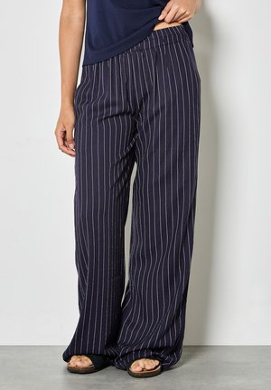 Person trägt marineblaue Wide-Leg-Hose mit weißen vertikalen Nadelstreifen und schwarze offene Pantoletten, steht vor einer hellen Wand.