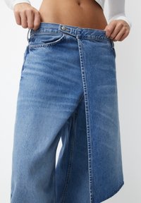 Culottes de mezclilla, de color azul claro, con una cinturilla alta y cierre de botón, y un diseño frontal abierto único. Textura suave con áreas desvaídas.
