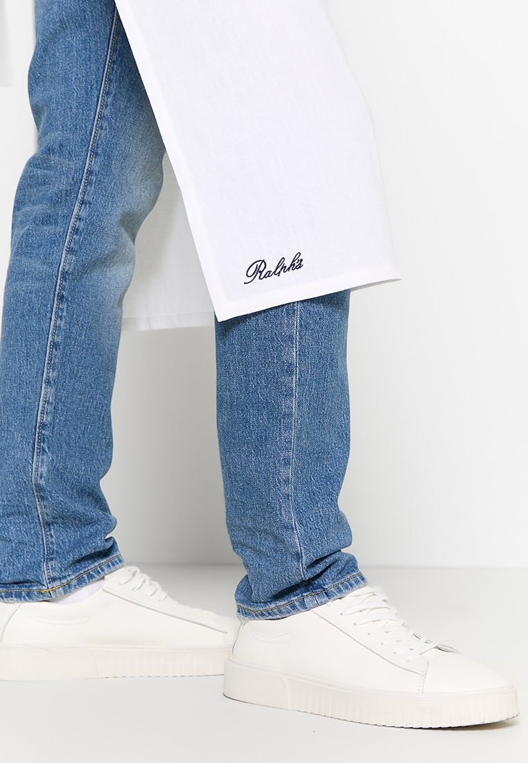 Grembiule bianco con testo ricamato blu marino, indossato sopra un paio di jeans di denim azzurri. Scarpe bianche con suola testurizzata e design minimale.