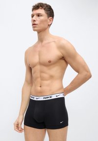 Άνδρας χωρίς πουκάμισο, με μαύρα boxer briefs Nike και λευκή μέση, στέκεται με το χέρι πίσω από την πλάτη, κοιτάζοντας προς το πλάι σε απλό φόντο.