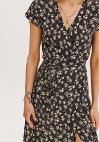 Čierna kvetinová šatka s krátkymi rukávmi, opaskom na viazanie a rozparkom. Ozdobená žltými a modrými kvetmi na tmavom pozadí.