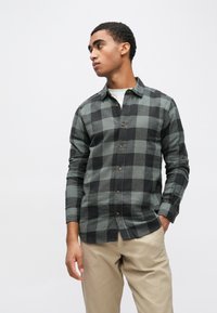Black and green plaid flannel shirt with a button-down front, long sleeves, and a rounded hem, paired with beige trousers.