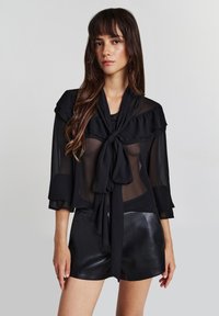 Schwarze, durchsichtige Bluse mit Rüschendetails und Halsband, mit Dreiviertelärmeln und kombiniert mit schwarzen Shorts.