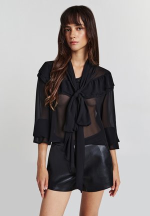 Blusa nera trasparente con dettagli arruffati e un fiocco al collo, con maniche a tre quarti e abbinata a pantaloni corti neri.