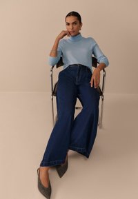 Lichtblauwe coltrui met lange mouwen, gecombineerd met wijde donkerblauwe denim jeans en gestreepte grijze puntige hakken. Zittend op een stoel.