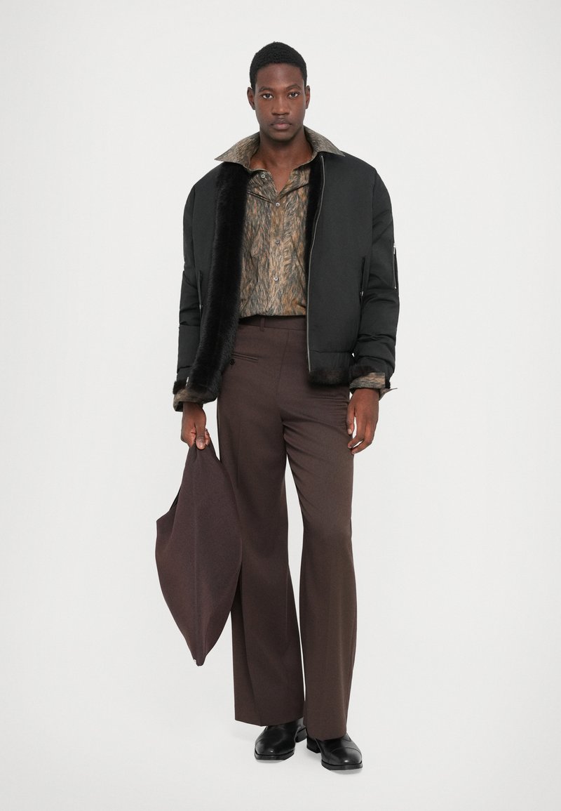 Giacca a vento nera con colletto in pelliccia sintetica, camicia con motivo in toni marroni, pantaloni marroni e borsa marrone scuro. Scarpe nere.