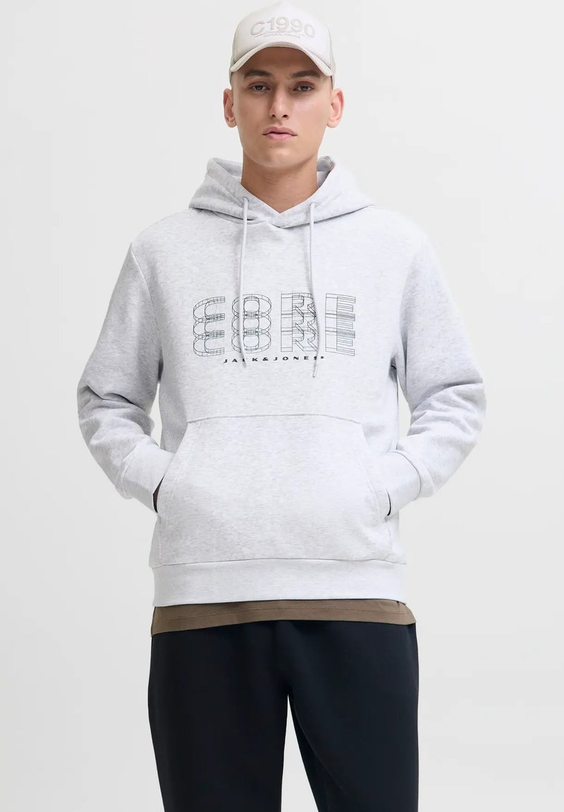 Giovane uomo che indossa una felpa leggera grigia con grafica "CORE", cappellino beige, pantaloni scuri e mani nelle tasche della felpa, contro uno sfondo neutro.