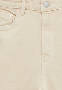Gros plan d’un jean en denim beige montrant les poches avant, la boucle de ceinture, les rivets métalliques, les coutures et un bouton en métal avec gravure de la marque.