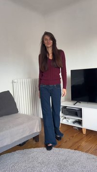 Bordowy sweter z prążkowanej dzianiny, ciemnoniebieskie dzwoniące dżinsy i czarne kapcie. Pokój wyposażony w szarą sofę, grzejnik i szafkę pod telewizor z elektroniką.