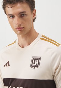 Maglia da calcio beige e marrone a maniche corte; presenta un colletto a girocollo, tre strisce dorate sulle spalle e un'emblema del club sul petto.