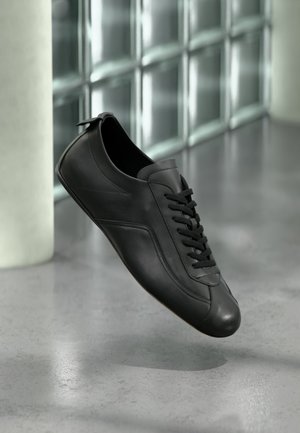 Zwarte leren sneaker met veters zwevend boven een gepolierde betonnen vloer naast een glasblokmuur in een minimalistische setting.