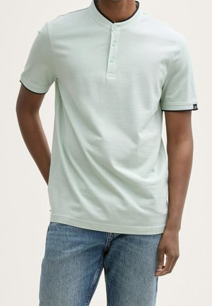 Mężczyzna w jasnozielonej koszulce henley z krótkim rękawem z trzema guzikami oraz niebieskich jeansach, stojący na tle jednolitego jasnego tła.