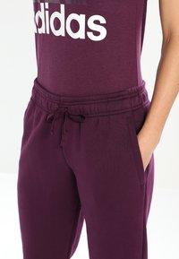 Lilla joggebukser med snøre i taljen, sidelommer og en afslappet pasform, parret med en tætsiddende lilla skjorte med et hvidt logo.