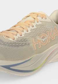 Μπεζ αθλητικό παπούτσι Hoka με κορδόνια ροδάκινου και λεπτομέρεια με ουράνιο τόξο στην σόλα, εμφανίζοντας το λογότυπο Hoka στο πλάι.
