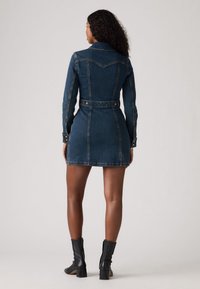 Vestito camicia in denim blu scuro con vita aderente, maniche lunghe e bottoni a pressione. Presenta una gonna svasata e cuciture a contrasto.
