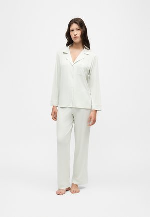 Jeune femme debout pieds nus, portant un haut blanc boutonné à manches longues et un pantalon ample assorti sur un fond blanc uni.