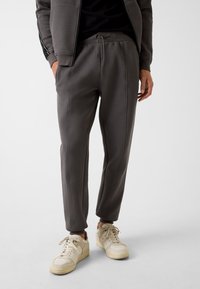 Dunkelgraue Jogginghose mit Kordelzugbund, Seitentaschen, elastischen Bündchen und kontrastierenden Steppnähten, kombiniert mit hellen Sneakers.