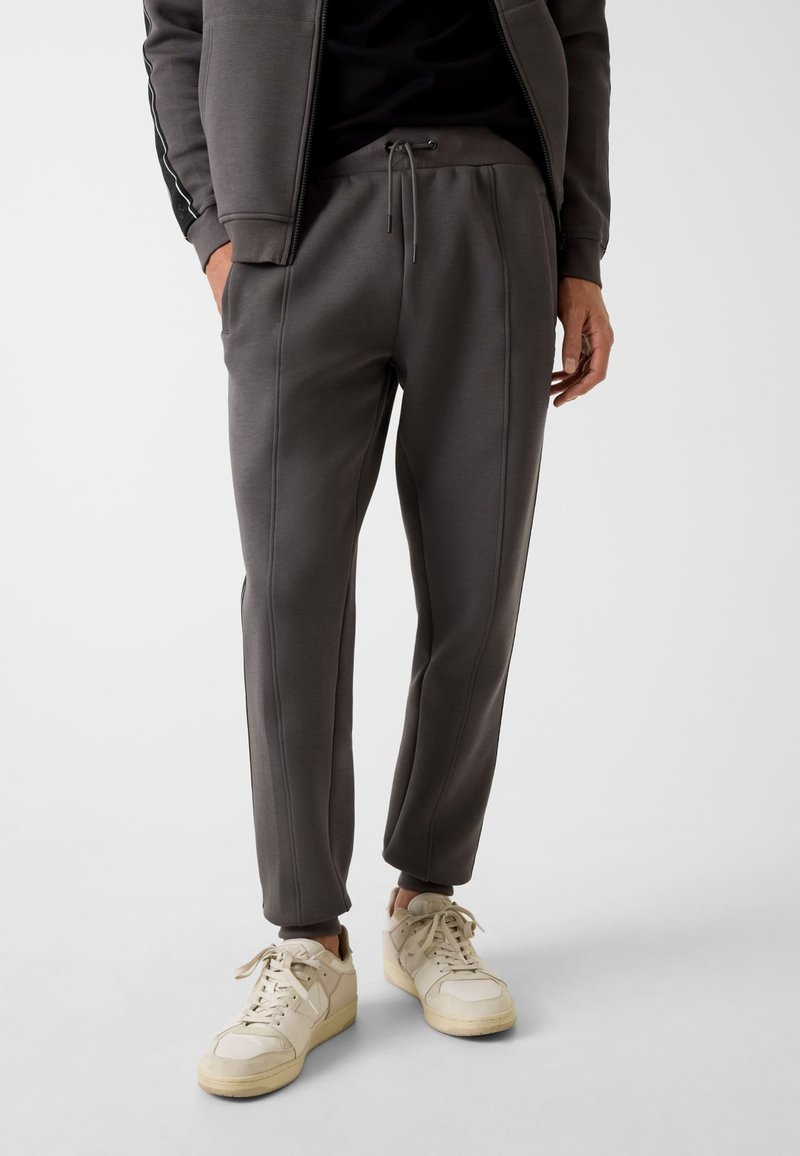 Dunkelgraue Jogginghose mit Kordelzugbund, Seitentaschen, elastischen Bündchen und kontrastierenden Steppnähten, kombiniert mit hellen Sneakers.