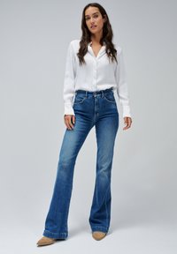 Jeans di denim blu a campana con un'alta vita, abbinati a una camicia bianca a maniche lunghe con bottoni. Il materiale è liscio e ha una vestibilità informale.