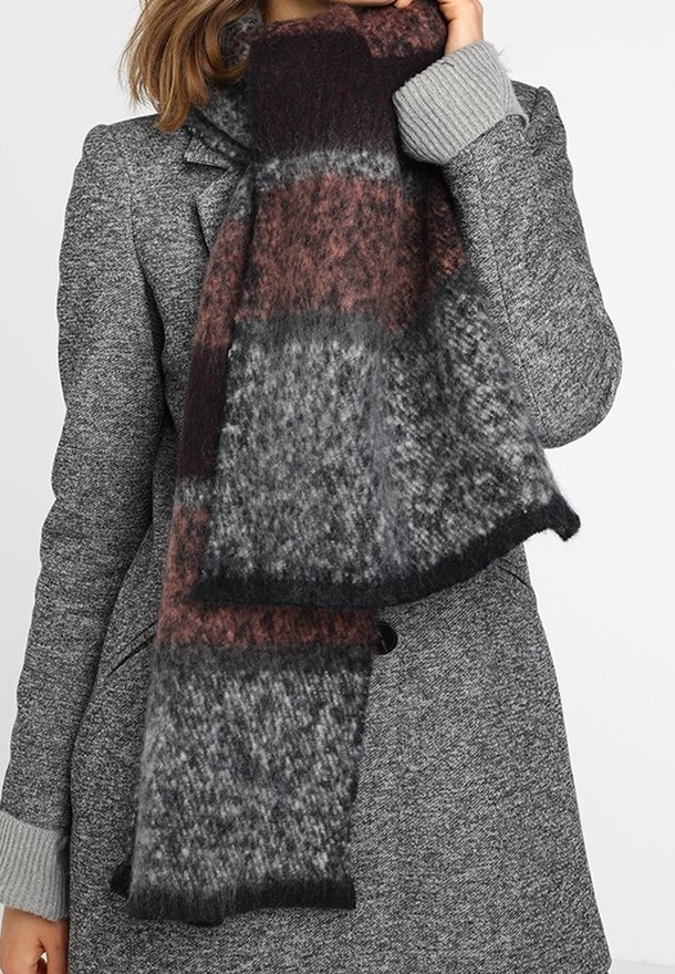 Femme portant un manteau gris texturé avec des poignets côtelés et une écharpe épaisse et duveteuse en motif patchwork noir, gris et rouge foncé.