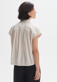 Light beige, short-sleeved blouse with a gathered back and elastic hem. Smooth fabric with a collar and minimal stitching details.