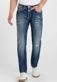 Jeans de mezclilla azul para hombre con un corte recto, detalles ligeramente desgastados y un diseño clásico de cinco bolsillos, combinados con zapatillas blancas.