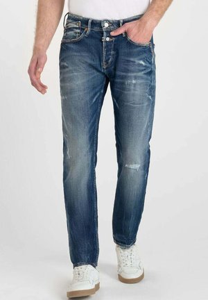 Jeans de mezclilla azul para hombre con un corte recto, detalles ligeramente desgastados y un diseño clásico de cinco bolsillos, combinados con zapatillas blancas.