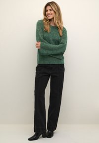 Grön texturerad tröja med långa ärmar, tillsammans med högmidjade svarta jeans och klackiga ankelstövlar. Enkel, modern design.