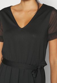 Černé šaty s výstřihem do V, krátkými průhlednými rukávy a úzkým pasem s vázacími šňůrkami. Hladká látka s mírným leskem a bez viditelných vzorů.