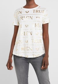 Žena v bílém tričku s krátkým rukávem a zlatým nápisem "TRUE RLGN", v kombinaci s vybledlými šedými džíny, stojí proti jednobarevnému pozadí.