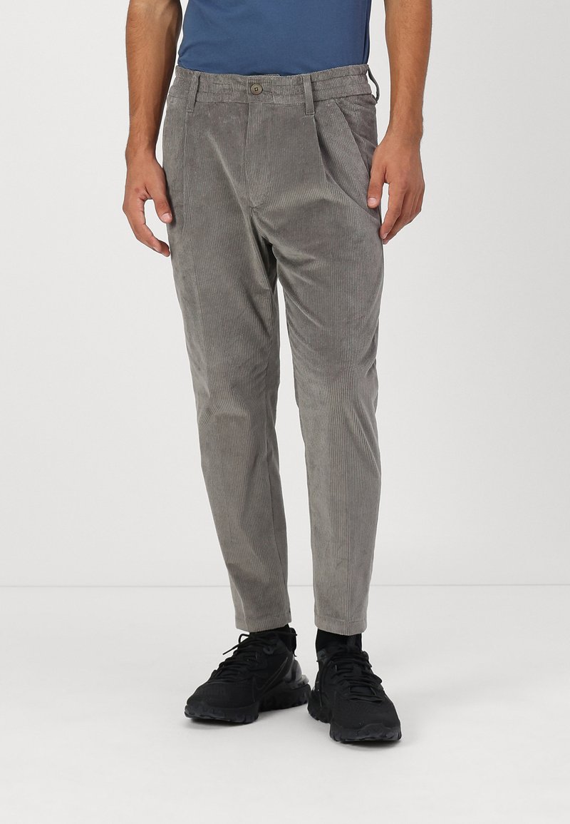 Pantalons en velours côtelé gris avec une finition texturée, dotés de plis et d'une fermeture à bouton. Associés à des chaussures de sport noires.