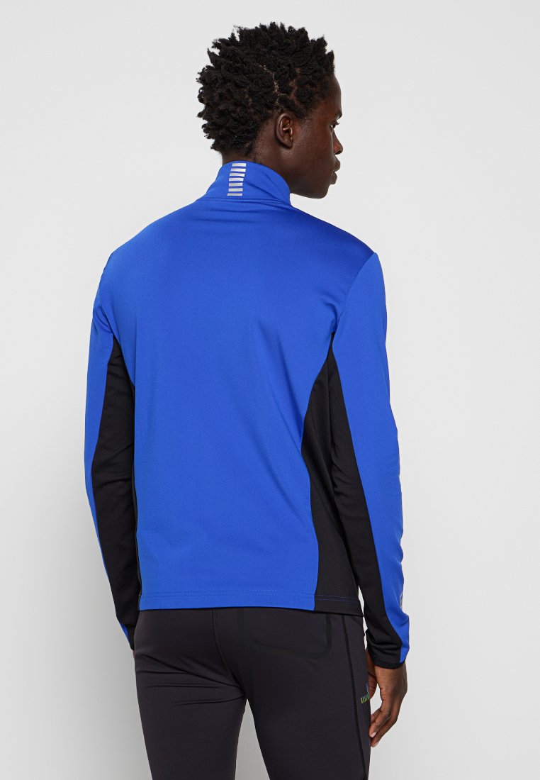Giacca sportiva blu con lati neri, colletto alto e dettagli riflettenti sulla parte posteriore. Realizzata in materiale liscio ed elastico.