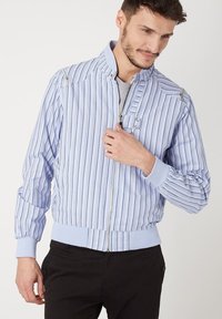 Chaqueta bomber a rayas azul y blanca con cremallera frontal, puños de punto y sutiles acentos metálicos en los hombros. Textura de tela suave.