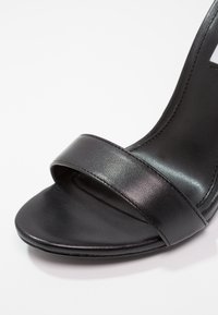 Gros plan sur l’avant d’une sandale en cuir noir avec une seule large bride sur les orteils et une semelle intérieure lisse.
