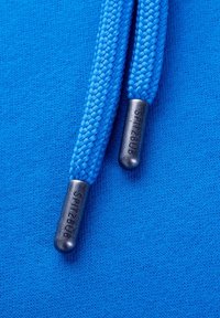 Gros plan de deux cordons de capuche tressés bleus avec des embouts en plastique noir embossés du mot Spitzbub sur un fond en tissu bleu.