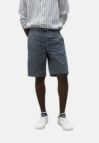 Pantalones cortos ligeros grises con cintura ajustable, bolsillos laterales y un corte relajado, combinados con una camisa de botones a rayas y zapatillas blancas.