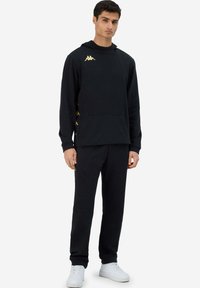 Completo con felpa nera e pantaloni della tuta realizzato in tessuto morbido, con tasca frontale e logo dorato sul petto e dettagli laterali. Sneakers bianche.